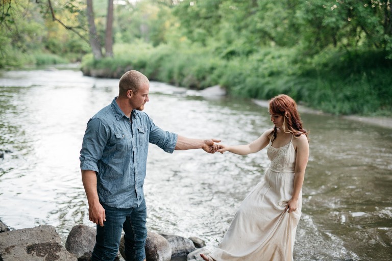 man helping his girlfriend walk on river rocks while she holds her dress
