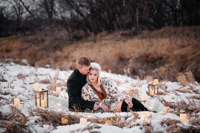 man and woman cuddling on white fur rug in the snow surrounded by candles