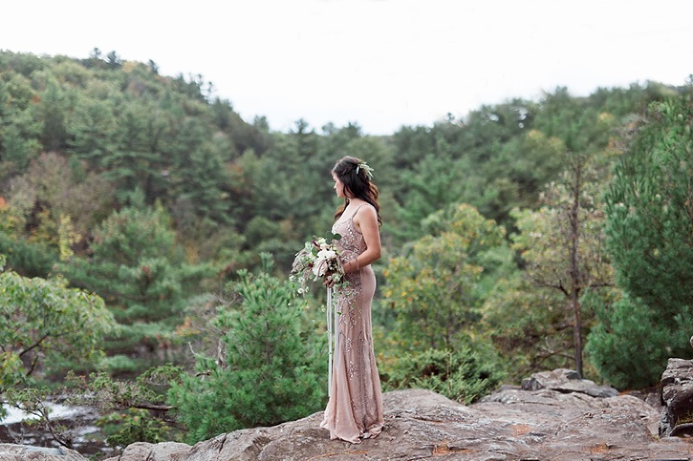 Bride on cliff in beaded gown, blue skirt, and shawl with bouquet